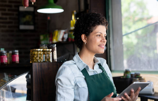 Business owner of a charcuterie looking away day dreaming and smiling while holding a tablet  - Business owner and place of work concepts