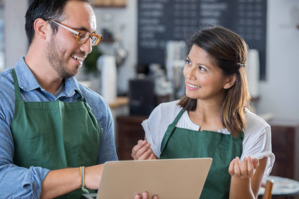 Confident male coffee shop manager trains a new female employee. They are looking at something on a laptop.